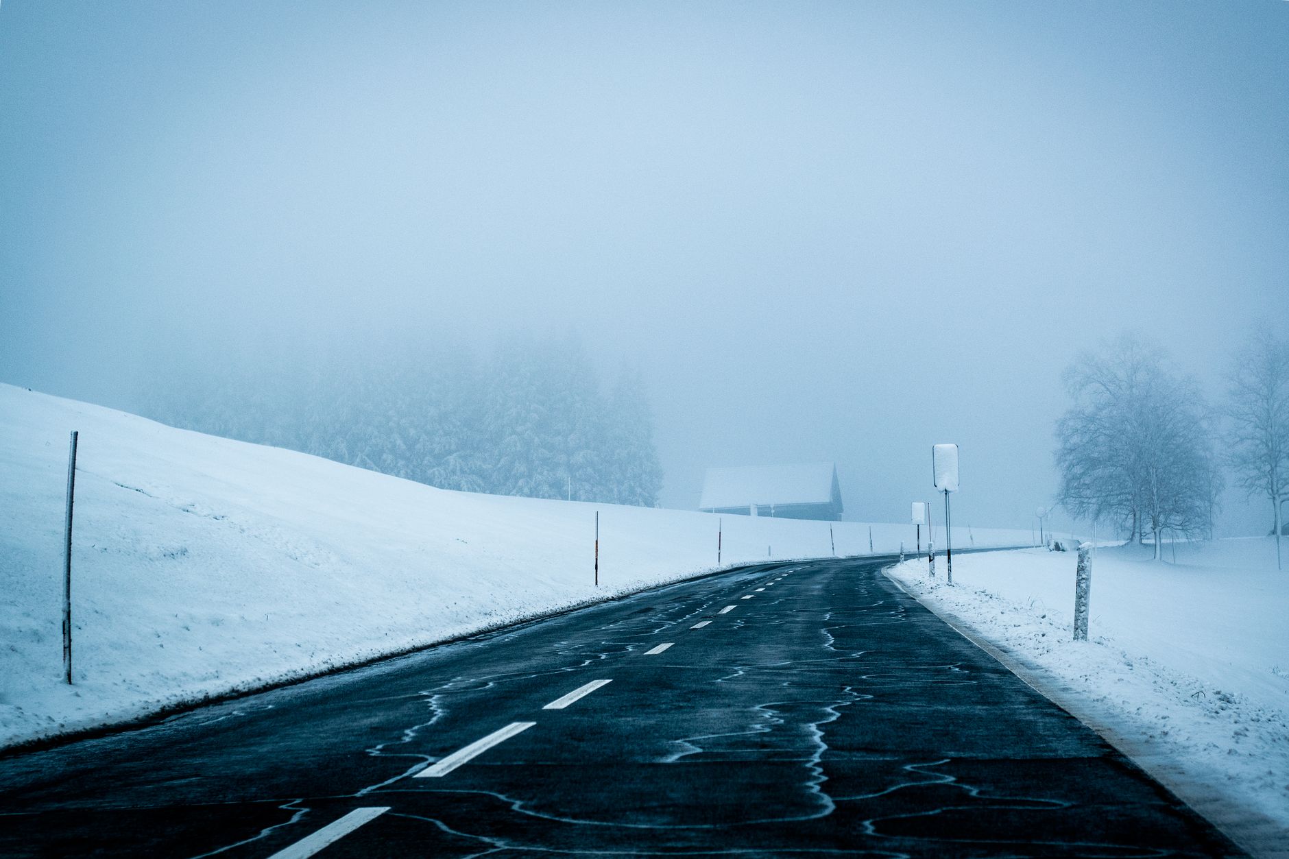 A foggy, snow-covered road stretches through a serene winter landscape, enveloped in mist.