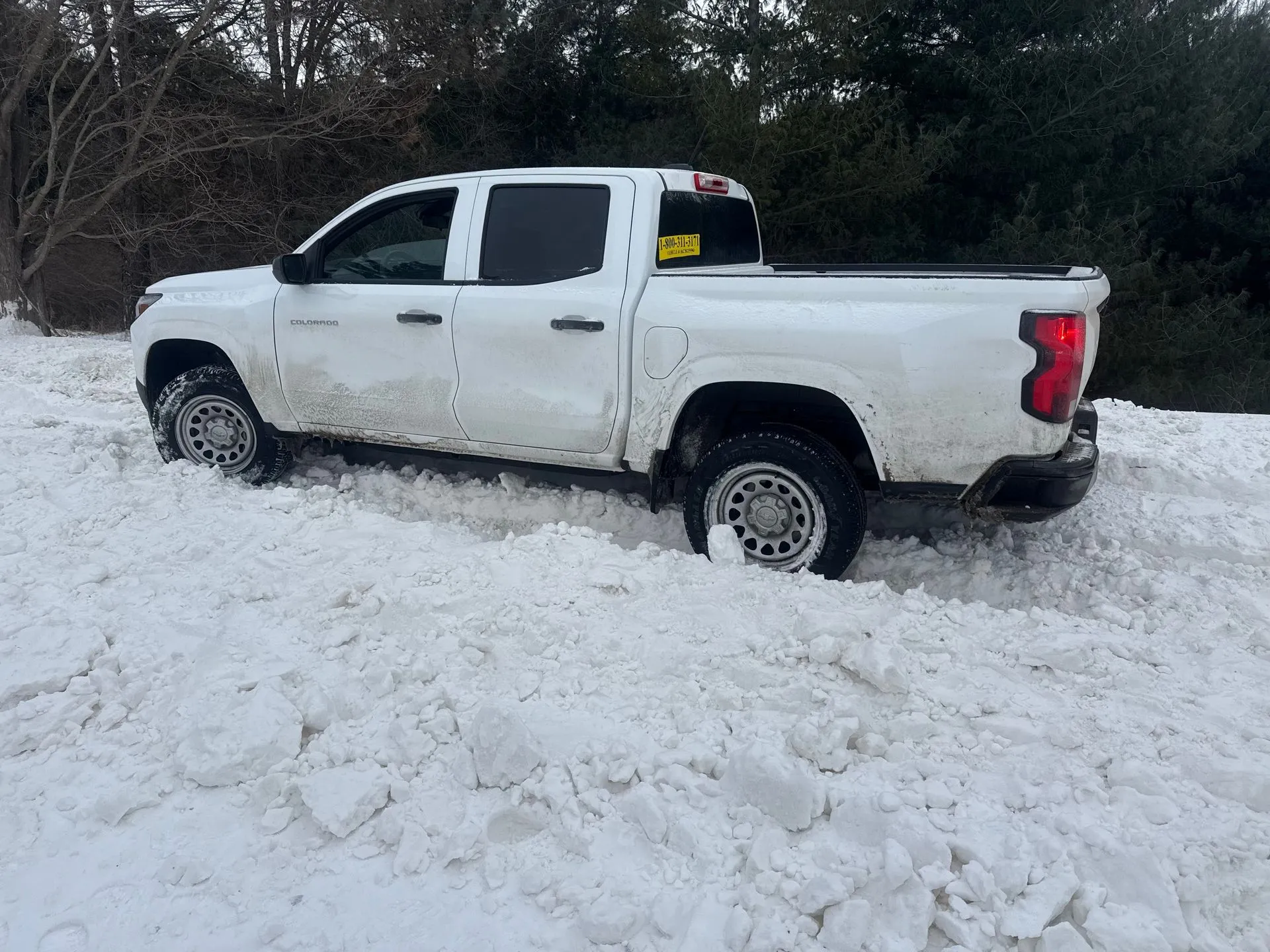 A white pickup truck stuck in a deep snowbank in a wooded area.