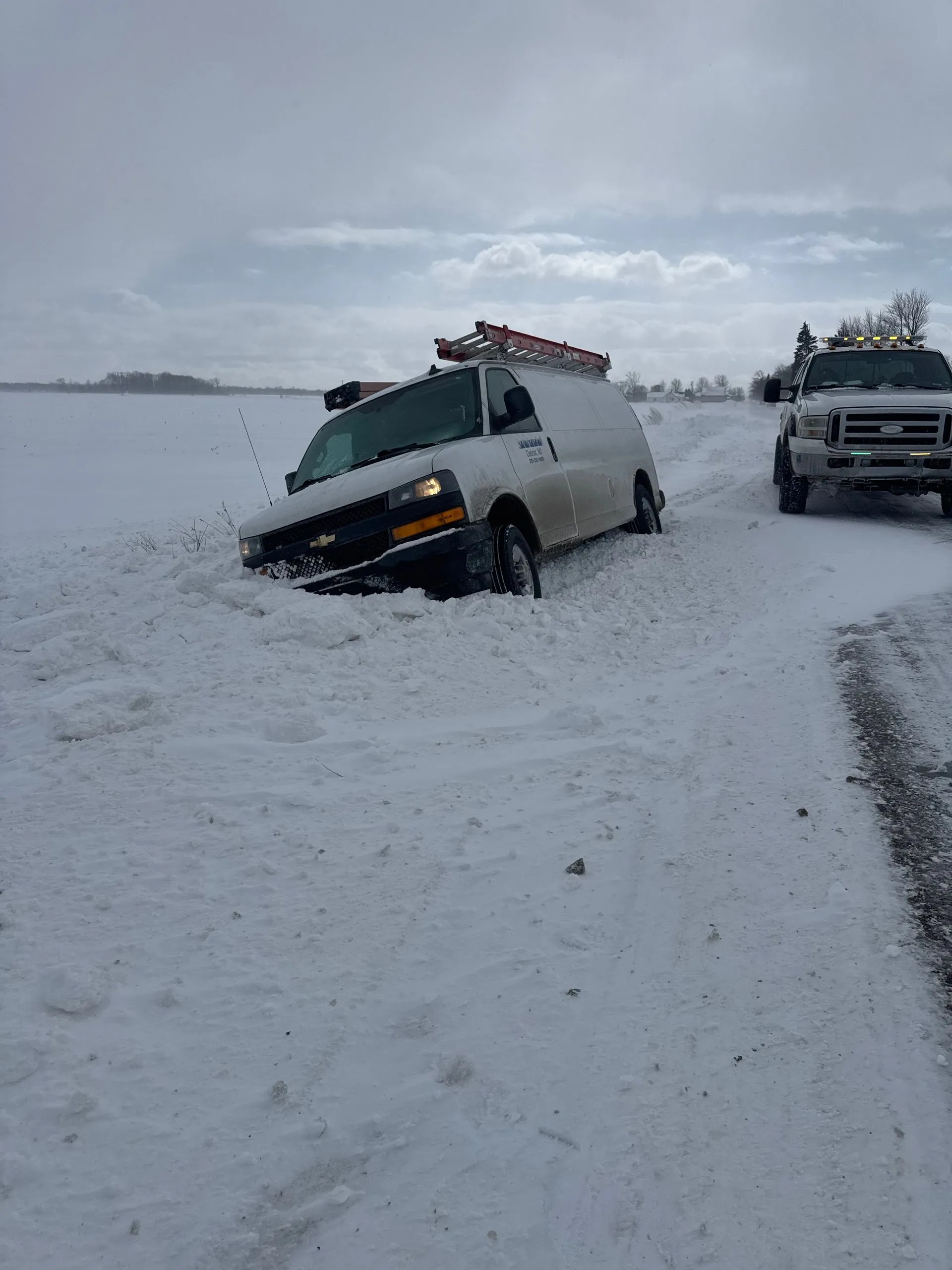 A white work van stuck in a snowbank on a snowy, open landscape with a support truck parked nearby.