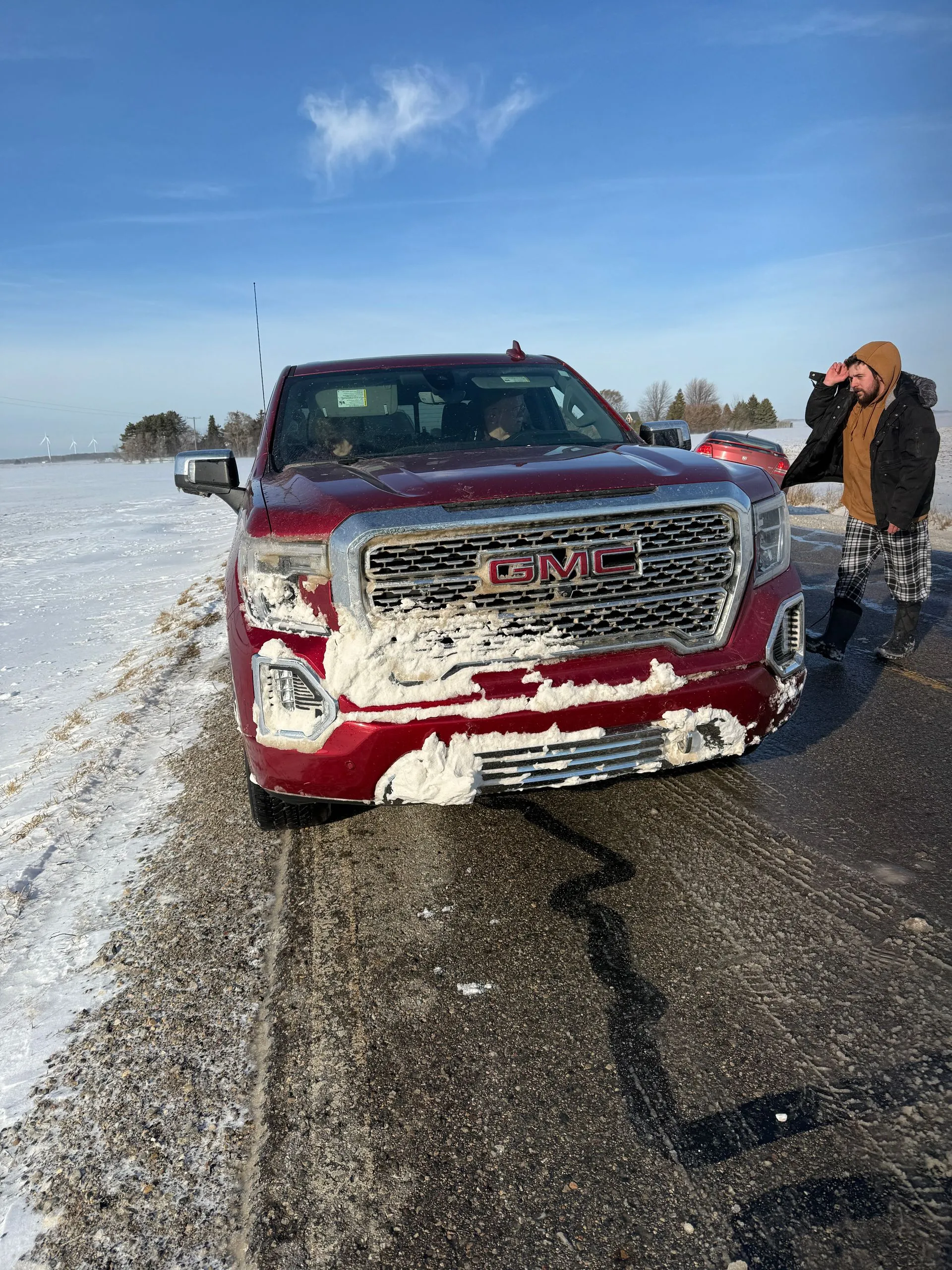 A man is standing next to a red gmc truck covered in snow.