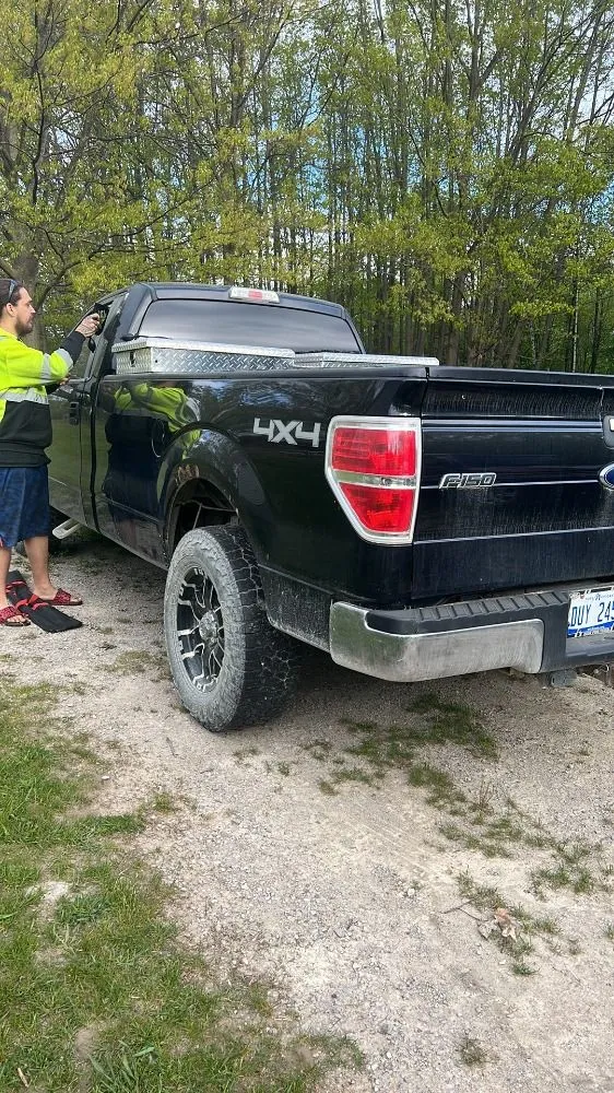 A person in a high-visibility yellow jacket stands beside a black Ford pickup truck on a gravel path near trees.